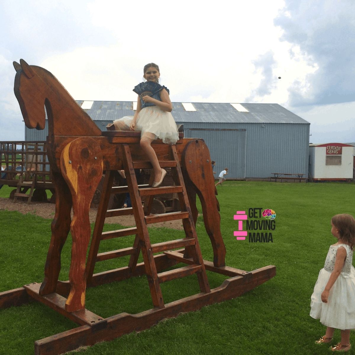 A picture of a girl sitting on a giant rocking horse at an outdoor wedding.
